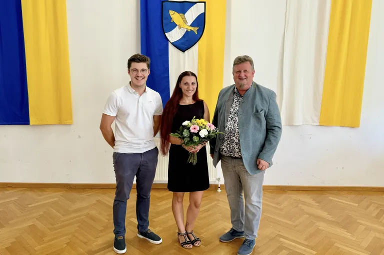 Drei Personen stehen vor einer Flagge. Die Frau hält einen Blumenstrauß.