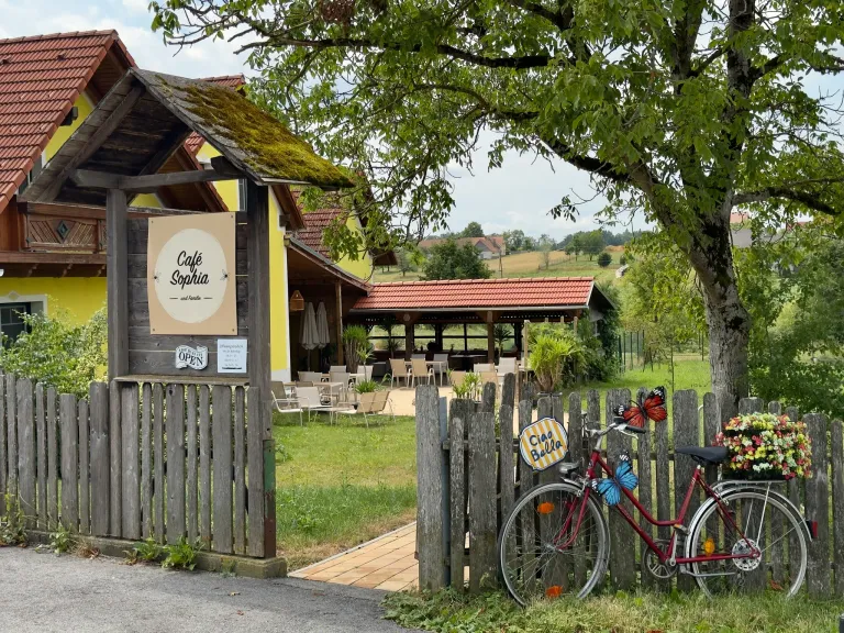 Cafe Sophia, ein charmantes Outdoor-Cafe mit einem Holzzaun, einem roten Fahrrad mit Schmetterlingsdekorationen und einem Schild, das anzeigt, dass es geöffnet ist.