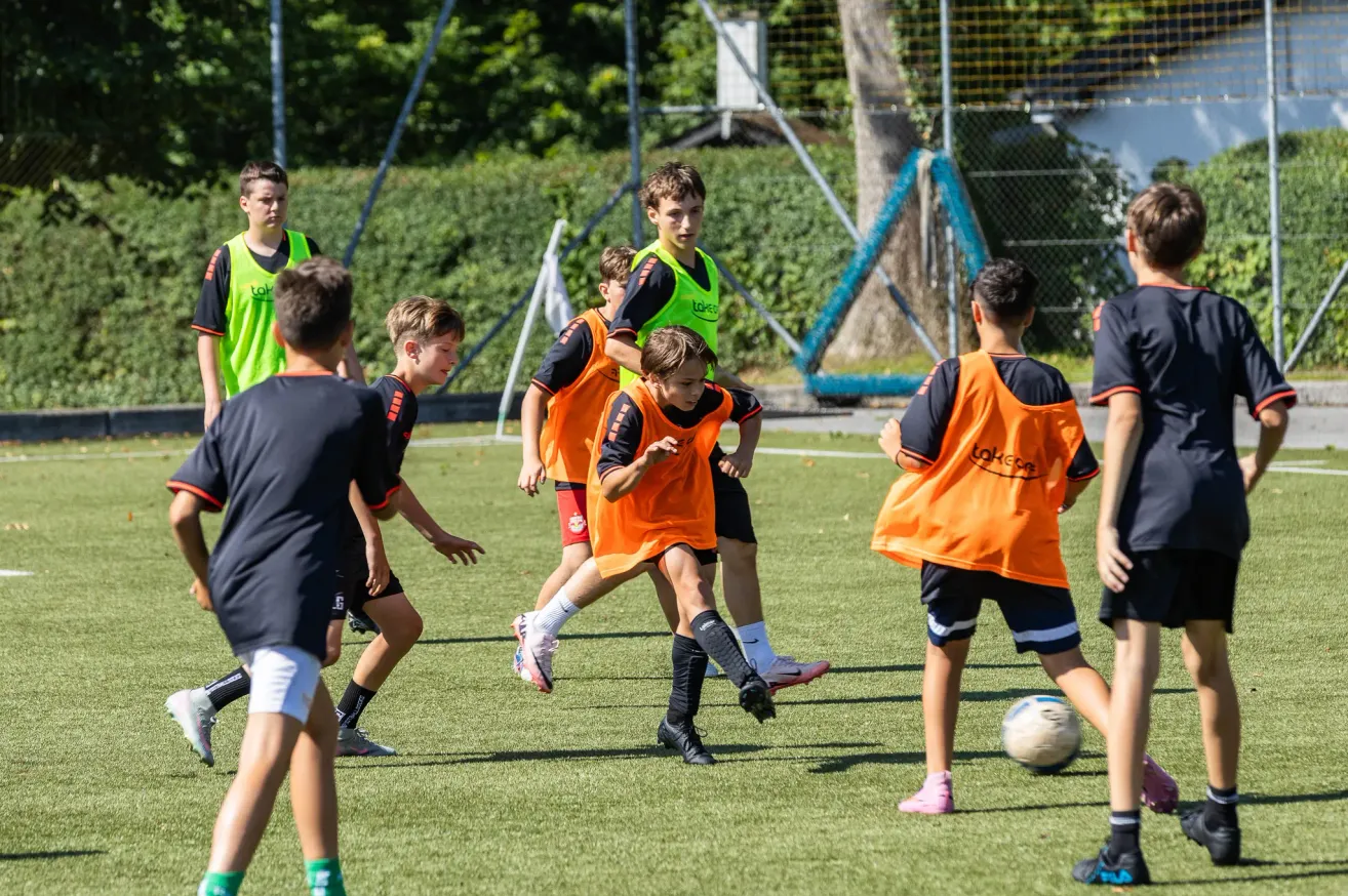 Eine Gruppe junger Jungen spielt Fußball auf einem Feld. Sie tragen orange Westen und schwarze Shorts. Ein Junge läuft auf den Ball zu, während andere ihn anschauen.