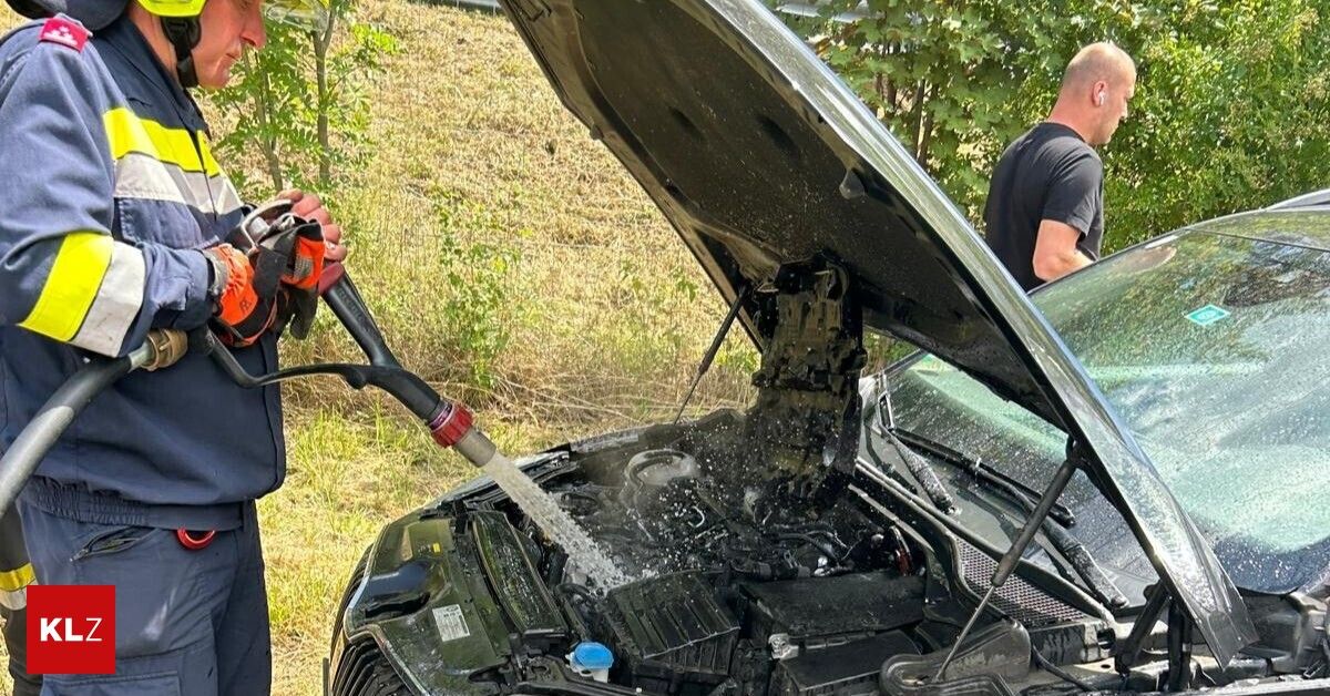 Eine Person reinigt den Motorraum eines Autos mit einem Schlauch, wobei die Motorhaube offen ist und Wasser auf den Motor spritzt.