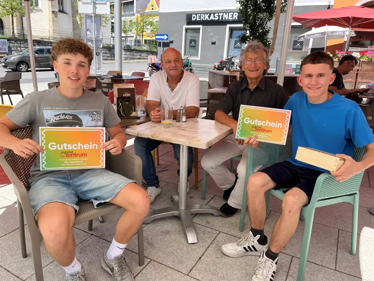 Vier Personen sitzen um einen Tisch in einem Café im Freien. Zwei junge Männer halten Gutscheine hoch, während zwei ältere Männer hinter ihnen sitzen.