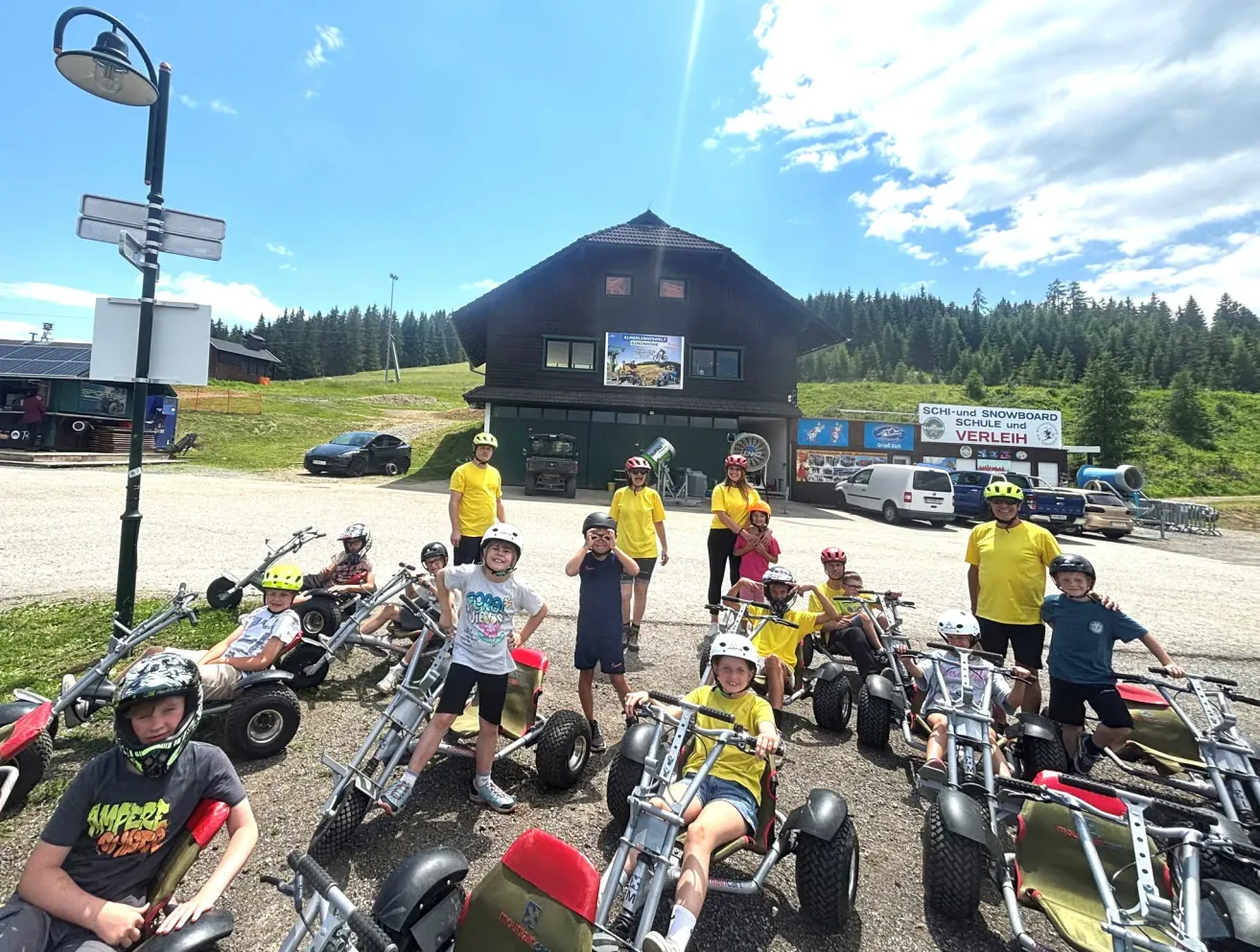 Eine Gruppe von Kindern und Erwachsenen in gelben Hemden und Helmen posiert für ein Foto vor einem Gebäude mit einem Schild, das 'Schi-und Snowboard Schule und Verleih' steht. Sie sitzen auf Go-Karts auf einem Parkplatz mit Bäumen und einem Auto im Hintergrund.