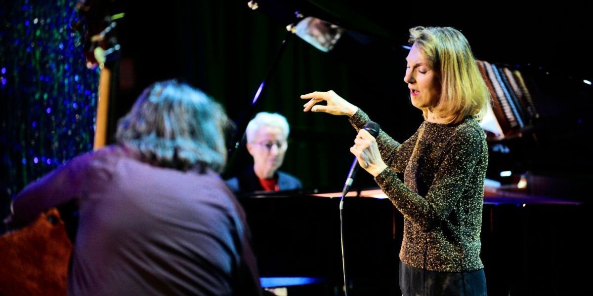 Eine Frau singt in ein Mikrofon auf der Bühne, mit einem Pianisten und einer anderen Person im Hintergrund.