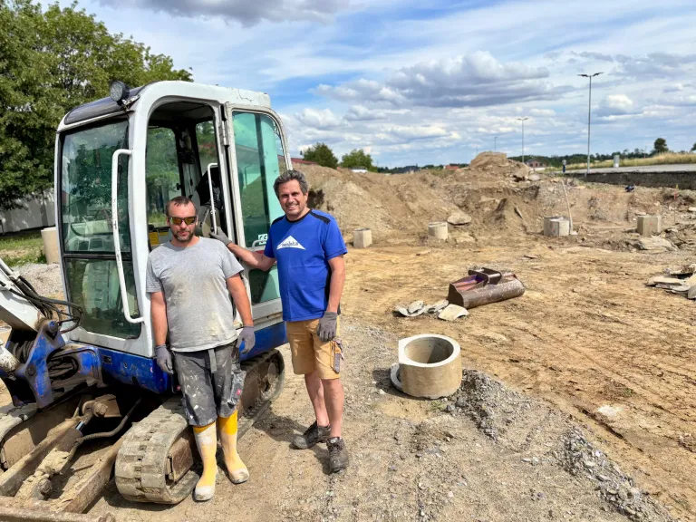 Zwei Männer in Arbeitskleidung stehen neben einem Baufahrzeug auf einer Baustelle, mit einem Haufen Erde und einer Betonröhre im Hintergrund.