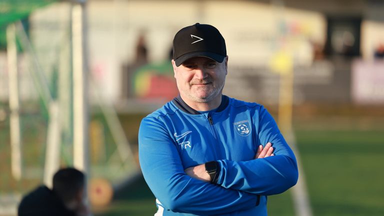 Ein Mann in einem blauen Jackett mit einer schwarzen Kappe und einem Nike-Logo lächelt für die Kamera auf einem Fußballfeld.