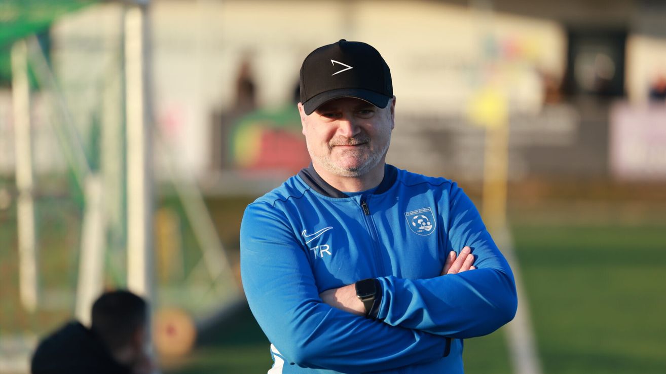 Ein Mann in einem blauen Jackett mit einer schwarzen Kappe und einem Nike-Logo lächelt für die Kamera auf einem Fußballfeld.