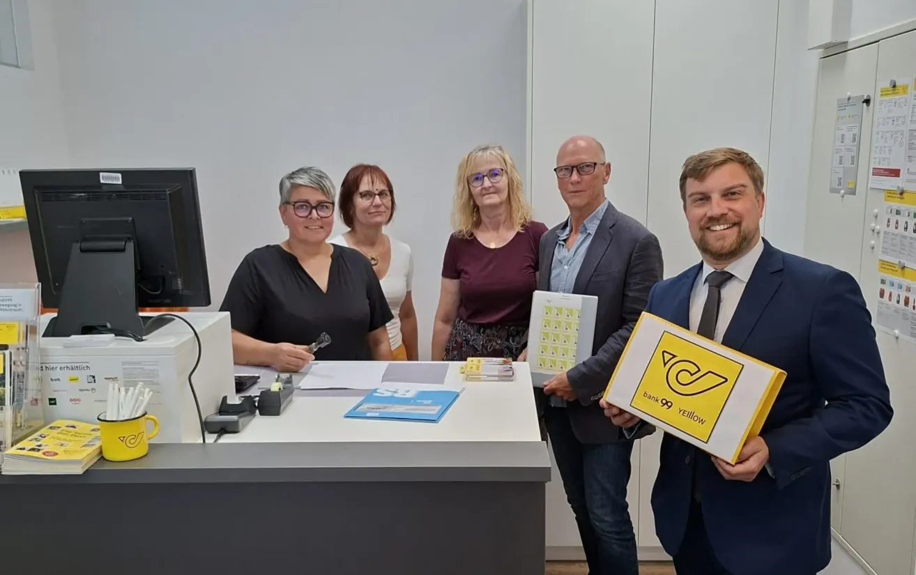 Fünf Personen stehen lächelnd hinter einem Schreibtisch für ein Foto. Eine Frau auf der linken Seite hält einen Stift und Papier. Ein Mann auf der rechten Seite hält eine Box mit einem gelben Logo.