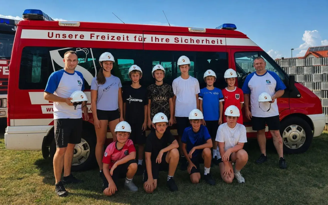 Eine Gruppe junger Menschen und Erwachsene posiert für ein Foto vor einem roten Feuerwehrwagen mit der Aufschrift 'Sere Freizeit für Ihre Sicherheit'. Sie tragen alle weiße Helme.