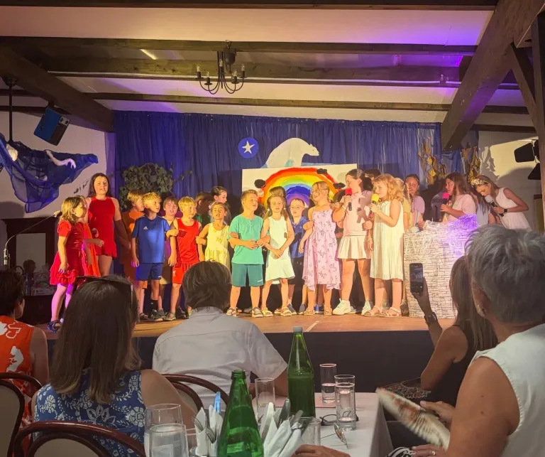 Eine Gruppe von Kindern in verschiedenen Farben steht auf einer Bühne mit einem Regenbogenhintergrund und singt in Mikrofone. Ein Publikum sitzt vor ihnen, und eine Frau hält ein Telefon.