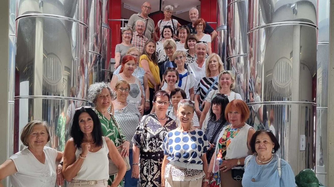 Eine Gruppe von Erwachsenen, hauptsächlich Frauen, lächelt und posiert für ein Foto vor einer metallischen Struktur. Sie tragen verschiedene Outfits und Accessoires.