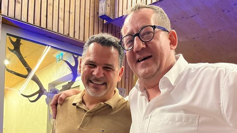 Zwei Männer lächeln und posieren für ein Foto, einer mit Bart und der andere mit Brille.