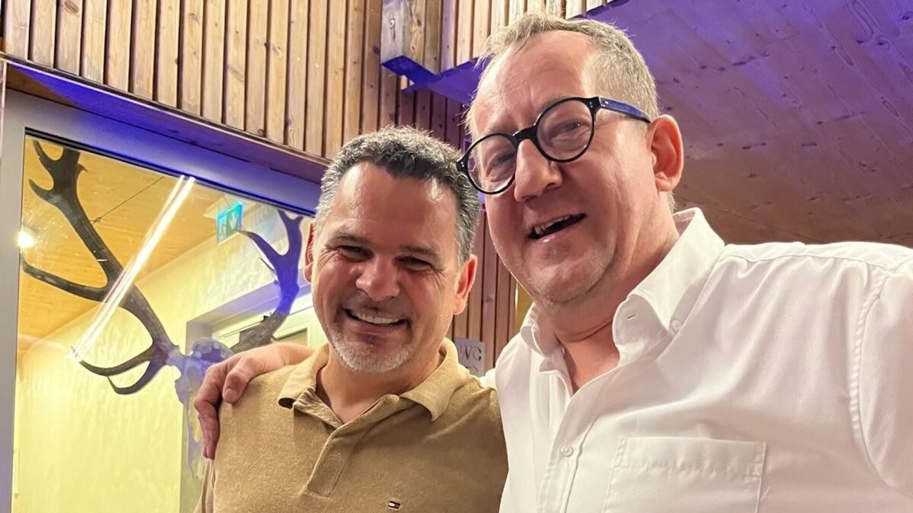 Zwei Männer lächeln und posieren für ein Foto, einer mit Bart und der andere mit Brille.