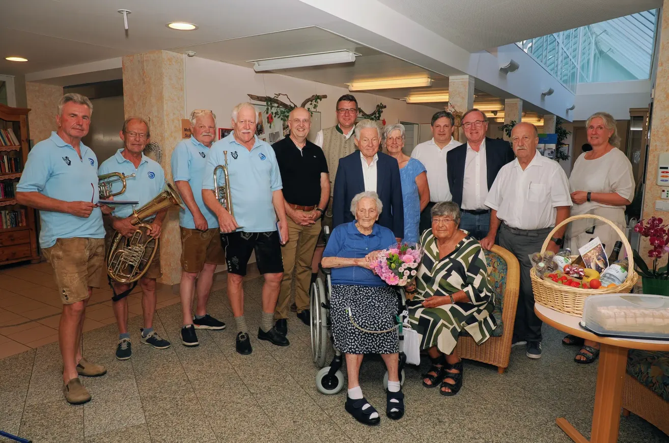 Eine Gruppe von Menschen, darunter zwei ältere Frauen, eine im Rollstuhl, posiert für ein Foto in einem gut beleuchteten Raum mit Musikinstrumenten und einem Korb mit Blumen.
