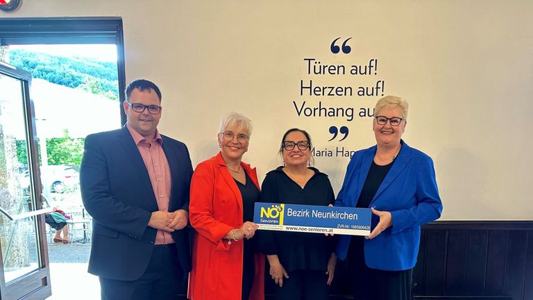 Vier Personen stehen zusammen vor einer Wand mit motivierenden Texten und halten ein blaues Schild mit 'NOES Senioren' und einer Website-Adresse.