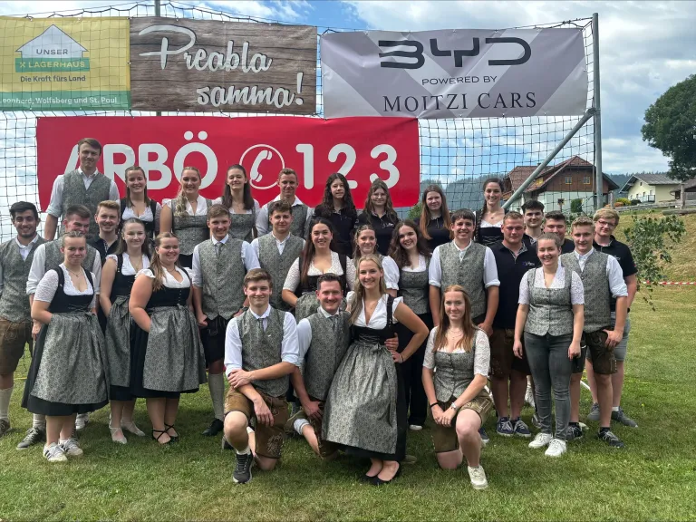 Eine Gruppe von Menschen in traditioneller bayerischer Kleidung posiert für ein Foto vor einem Banner mit der Nummer 123 und einem Logo.