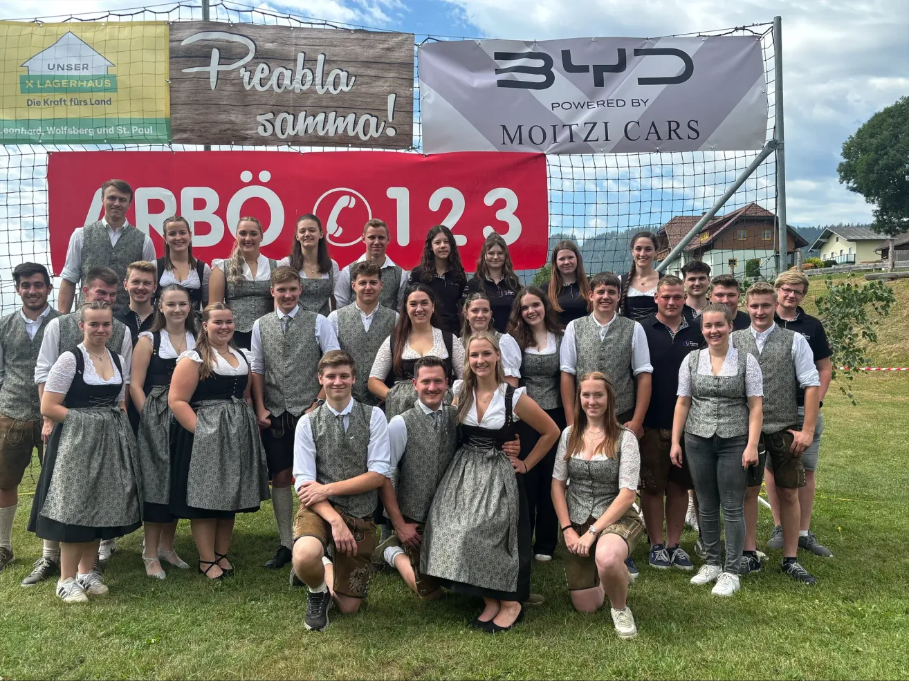 Eine Gruppe von Menschen in traditioneller bayerischer Kleidung posiert für ein Foto vor einem Banner mit der Nummer 123 und einem Logo.