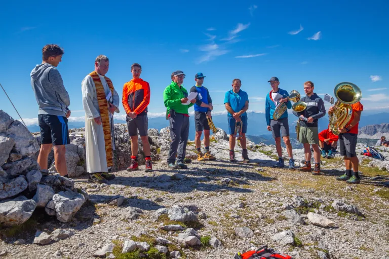 Eine Gruppe von Männern in Outdoor-Kleidung, einige halten Musikinstrumente, stehen auf einem felsigen Berggipfel unter einem klaren blauen Himmel.