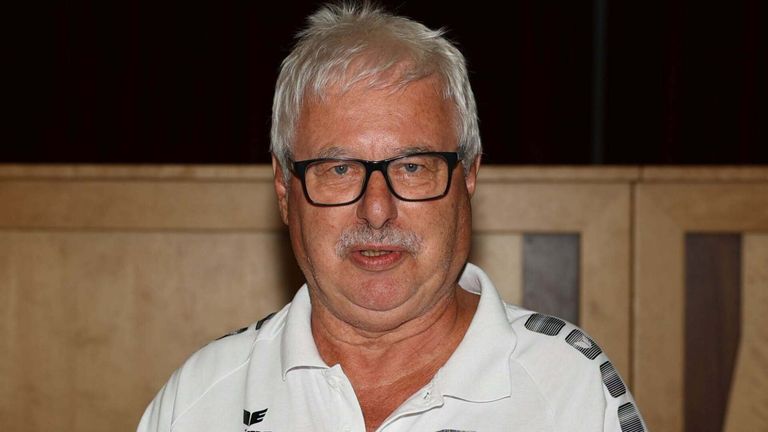 Ein älterer Mann mit weißem Haar und einem Schnurrbart, der eine Brille trägt und ein weißes Poloshirt mit einem Logo auf der linken Brustseite.