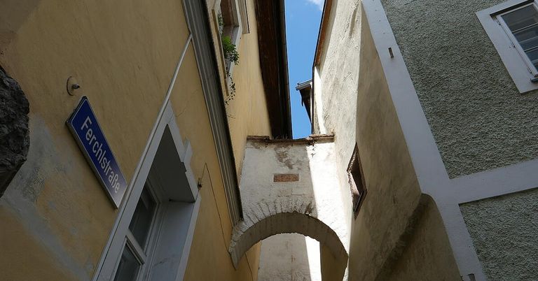 Eine schmale Gasse mit einem gelben Gebäude auf der linken Seite und einem weißen Bogen auf der rechten Seite, mit einem blauen Schild auf der linken Seite und einem Fenster auf der rechten Seite.