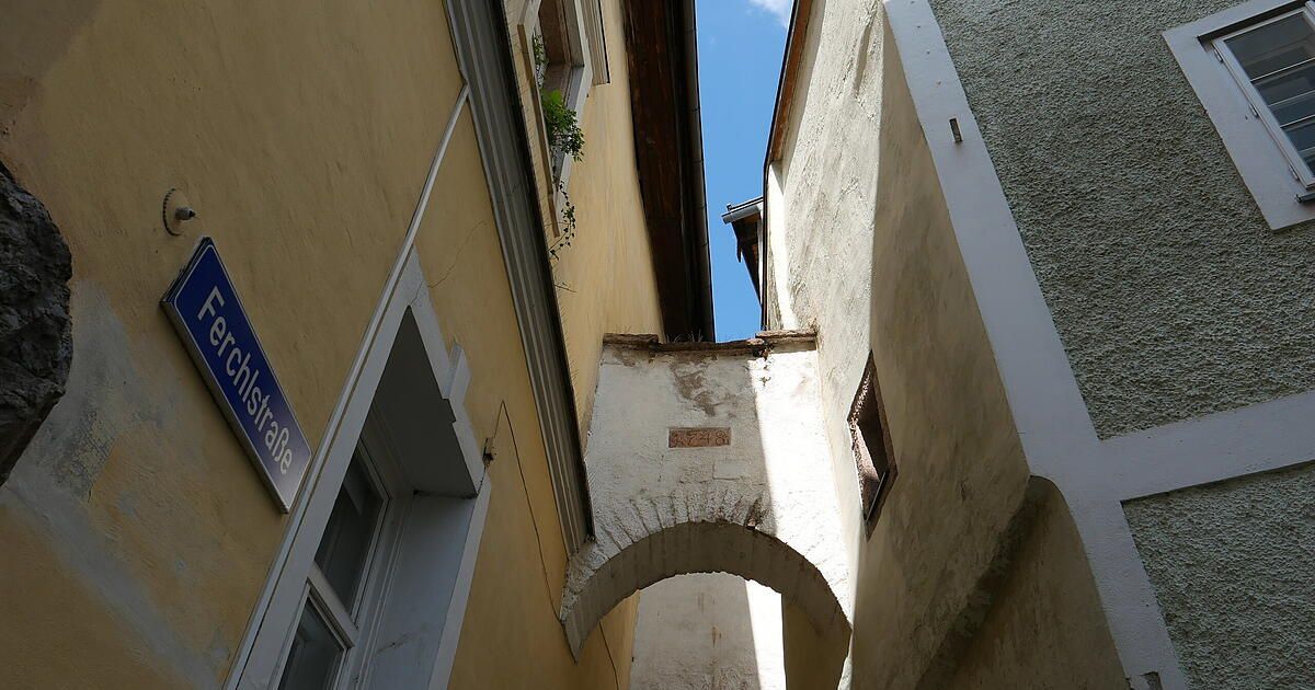 Eine schmale Gasse mit einem gelben Gebäude auf der linken Seite und einem weißen Bogen auf der rechten Seite, mit einem blauen Schild auf der linken Seite und einem Fenster auf der rechten Seite.