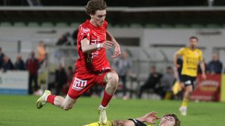 Ein Fußballspieler in einem roten Trikot mit dem Wort 'SCM' darauf läuft und springt in der Luft, möglicherweise versucht er, den Ball zu schießen. Ein anderer Spieler in einem gelben Trikot liegt auf dem Boden.