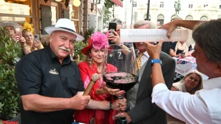 Ein Mann und eine Frau in formeller Kleidung halten ein großes Weinglas mit rotem Wein, während eine Person hinter ihnen ein Maßband hält. Sie stehen vor einem Tisch mit einer Flagge darauf.