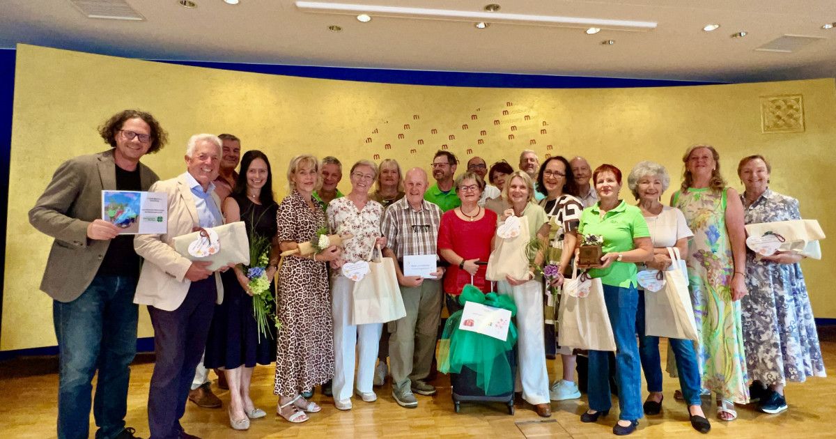 Eine Gruppe von Menschen, wahrscheinlich Freiwillige, steht zusammen und lächelt für ein Foto. Sie halten Blumen, Urkunden und Taschen. Die Wand hinter ihnen ist mit gelber Farbe und Zahlen dekoriert.