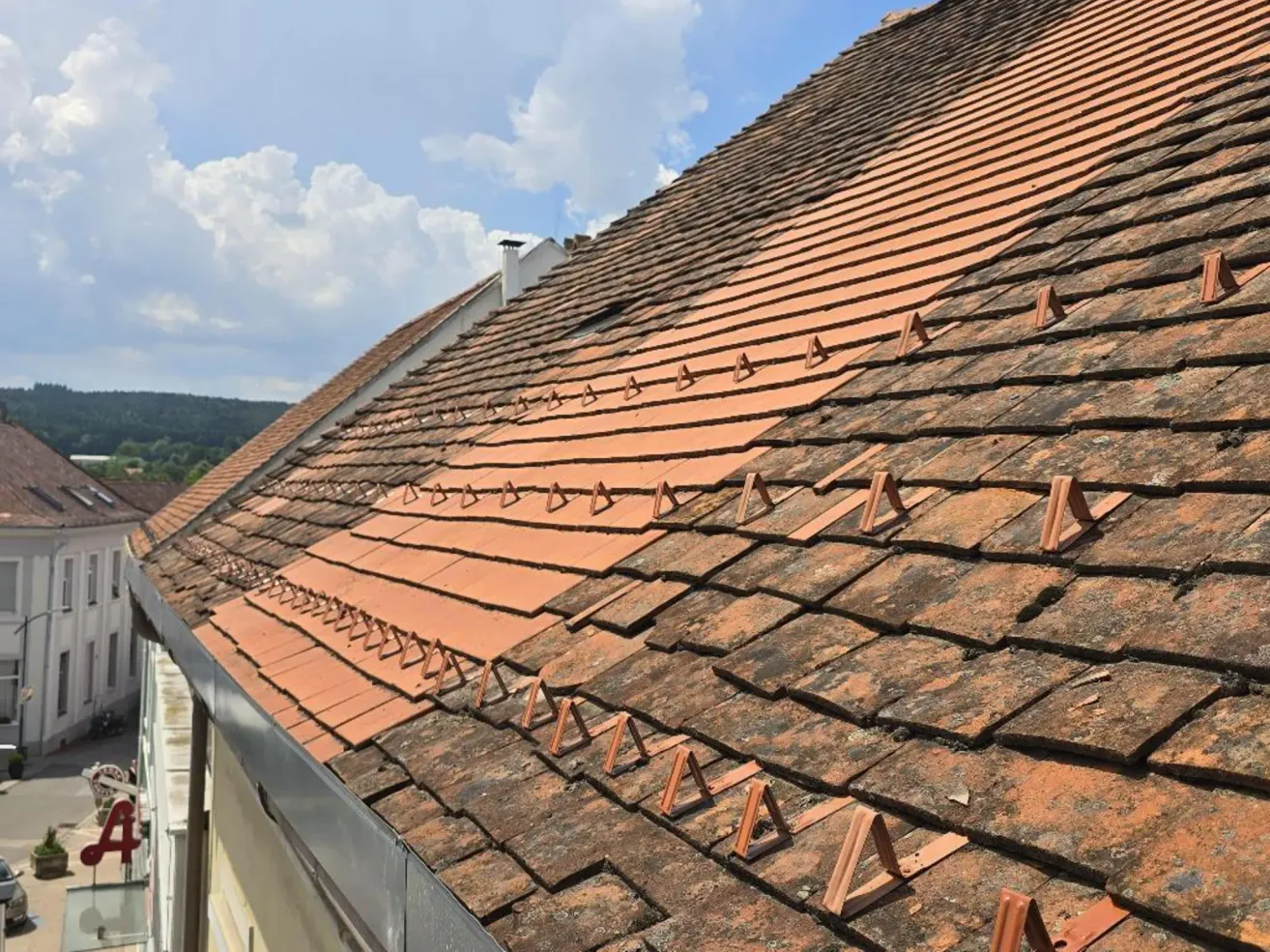 Eine Nahaufnahme eines roten Ziegeldachs mit dreieckigen Metallklammern, die die Ziegel halten. Der Himmel ist blau mit verstreuten Wolken.
