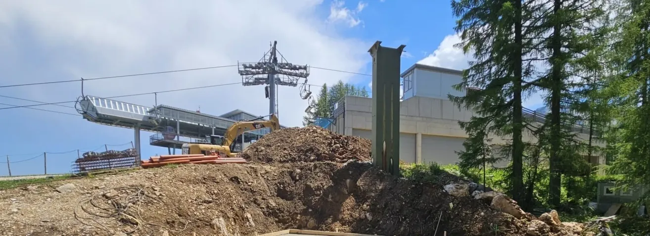 Eine Baustelle mit einem Haufen Erde, einem gelben Bagger und einer Skilift im Hintergrund.