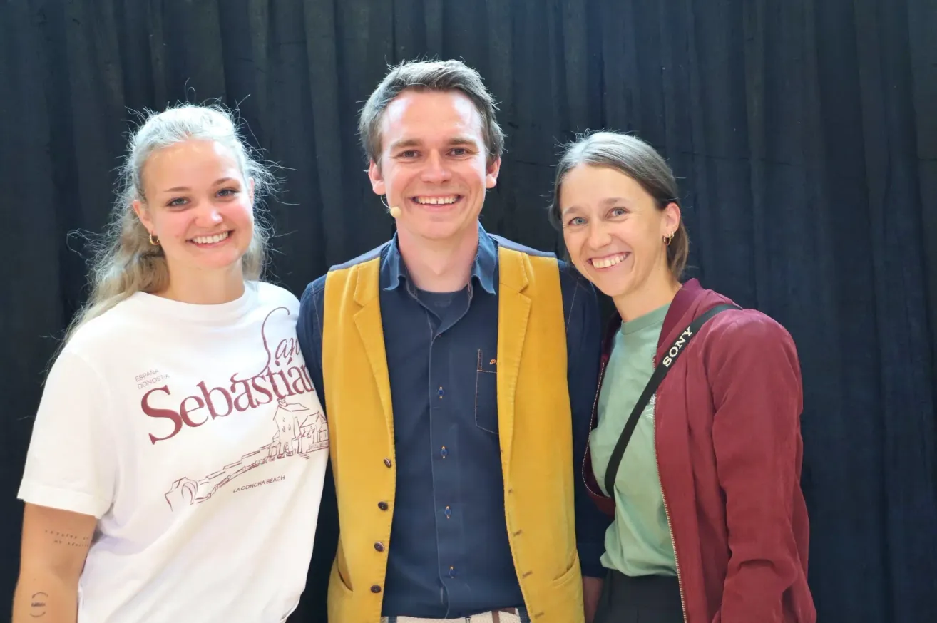 Drei Menschen lächeln und posieren für ein Foto. Der Mann in der Mitte hat ein Mikrofon am Ohr. Die Frau links trägt ein weißes T-Shirt mit dem Wort Sebastian. Die Frau rechts trägt eine kastanienbraune Jacke.