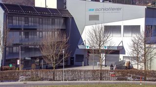 Ein großes Gebäude mit dem Namen Schloetterer an der Wand. Das Gebäude hat mehrere Fenster und eine Garagentür. Vor dem Gebäude stehen zwei Bäume. Eine Person steht in der Nähe der Treppe.