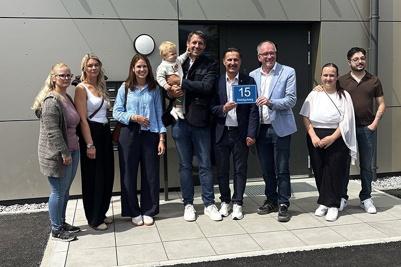 Eine Gruppe von Menschen steht vor einem Gebäude, lächelt und posiert für ein Foto. Ein Mann hält ein Baby und ein Schild mit der Nummer 15.