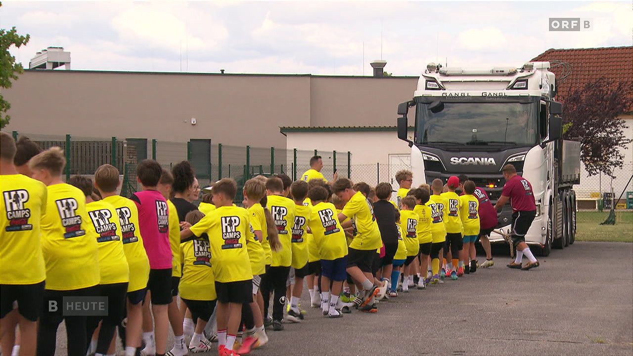 Eine Gruppe von Kindern in gelben T-Shirts mit der Aufschrift 'KP Camps' steht in einer Reihe, wahrscheinlich für eine Sportveranstaltung. Ein Scania-Lkw ist hinter ihnen geparkt.