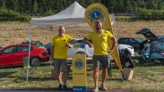 Zwei Männer in gelben Shirts stehen neben einem gelben Banner mit dem Lions Club Logo. Sie stehen vor einem Zelt und einer Spendenbox. Hinter ihnen sind mehrere Autos am Straßenrand geparkt.