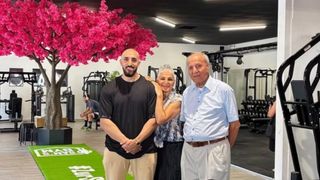 Drei Personen stehen in einem Fitnessstudio, wobei der Mann in der Mitte lächelt, während die Frau neben ihm seinen Schultern umschlungen hat. Dahinter steht ein Mann mit einem Lächeln. Im Hintergrund befinden sich Geräte und ein rosa Baum.