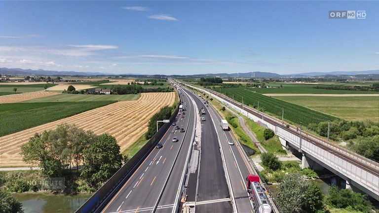 Luftaufnahme einer belebten Autobahn mit mehreren Spuren, Autos und Lastwagen, umgeben von üppigen grünen Feldern und Bäumen an einem sonnigen Tag.
