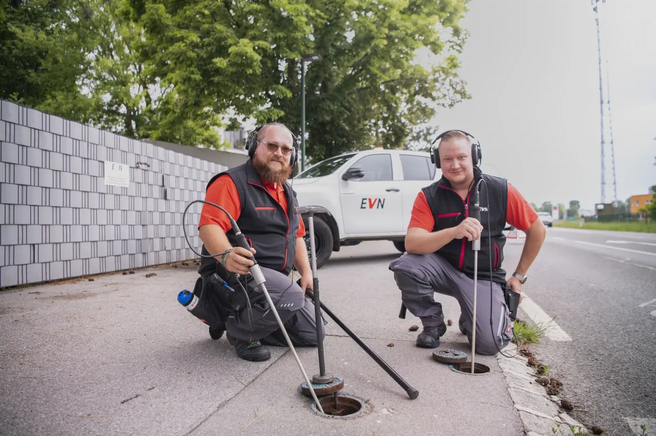 Zwei Männer in orangefarbenen Hemden und Kopfhörern knien neben einem Schacht, einer hält einen Metallstab und der andere ein Werkzeug. Ein weißer Lastwagen ist hinter ihnen geparkt.