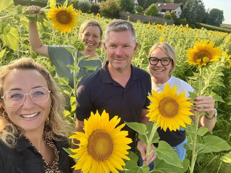 Vier Personen stehen in einem Sonnenblumenfeld, lächeln und posieren für ein Foto. Der Mann in der Mitte hat einen Bart und trägt ein dunkles Hemd. Die Frauen an seiner Seite tragen eine Brille und lächeln.