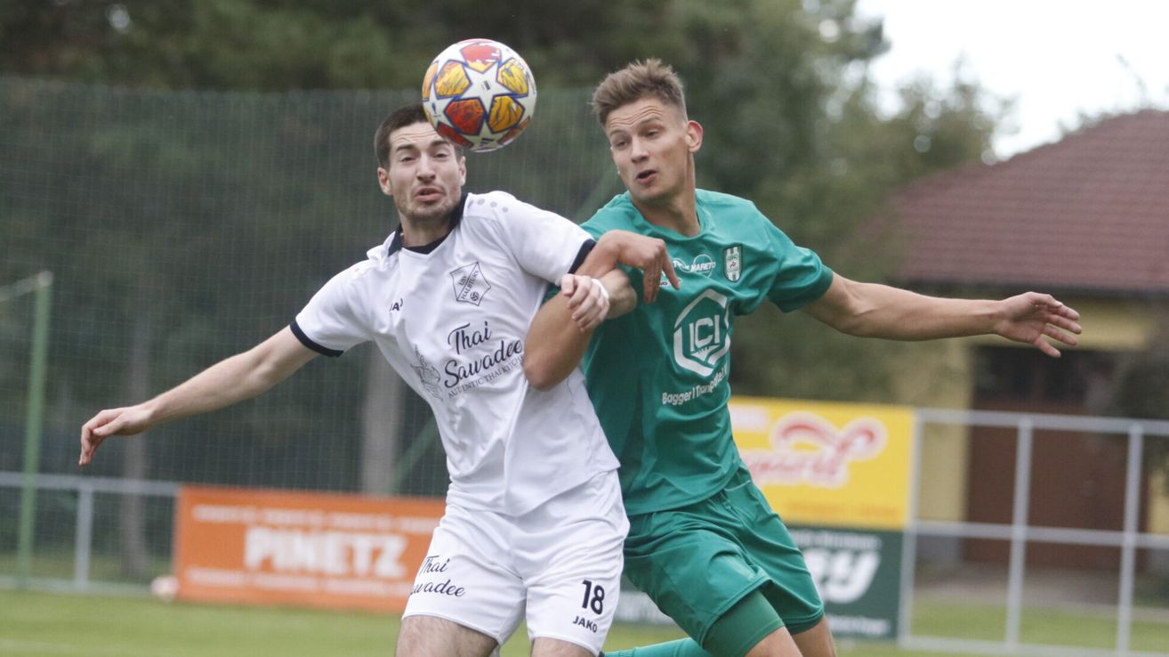 Zwei Fußballspieler, einer in einem weißen Trikot mit 'That Sawadee' und der Nummer 18, und der andere in einem grünen Trikot mit 'LCI', befinden sich in der Luft und konkurrieren um einen Fußball.