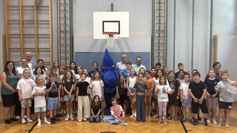 Eine Gruppe von Kindern und Erwachsenen posiert für ein Foto in einer Turnhalle mit einem Maskottchen in der Mitte. Das Maskottchen ist eine große blaue Kreatur. Die Turnhalle hat einen Basketballkorb und einen Holzboden.