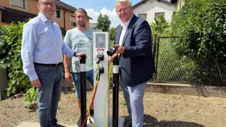 Drei Männer stehen neben einer Ladestation für Elektrofahrzeuge, wobei einer der Männer eine Kamera hält und die anderen beiden die Ladekabel halten. Sie lächeln und scheinen für ein Foto zu posieren.