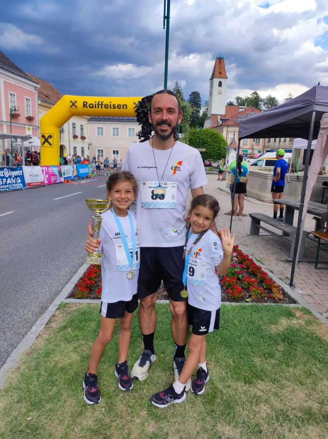 Ein Mann und zwei junge Mädchen stehen auf einem Rasen und posieren für ein Foto. Der Mann hält eine Trophäe und die Mädchen tragen Medaillen. Sie stehen vor einem gelben Bogen mit dem Wort Raiffeisen. Dahinter steht ein Zelt, eine Bank und ein Gebäude.