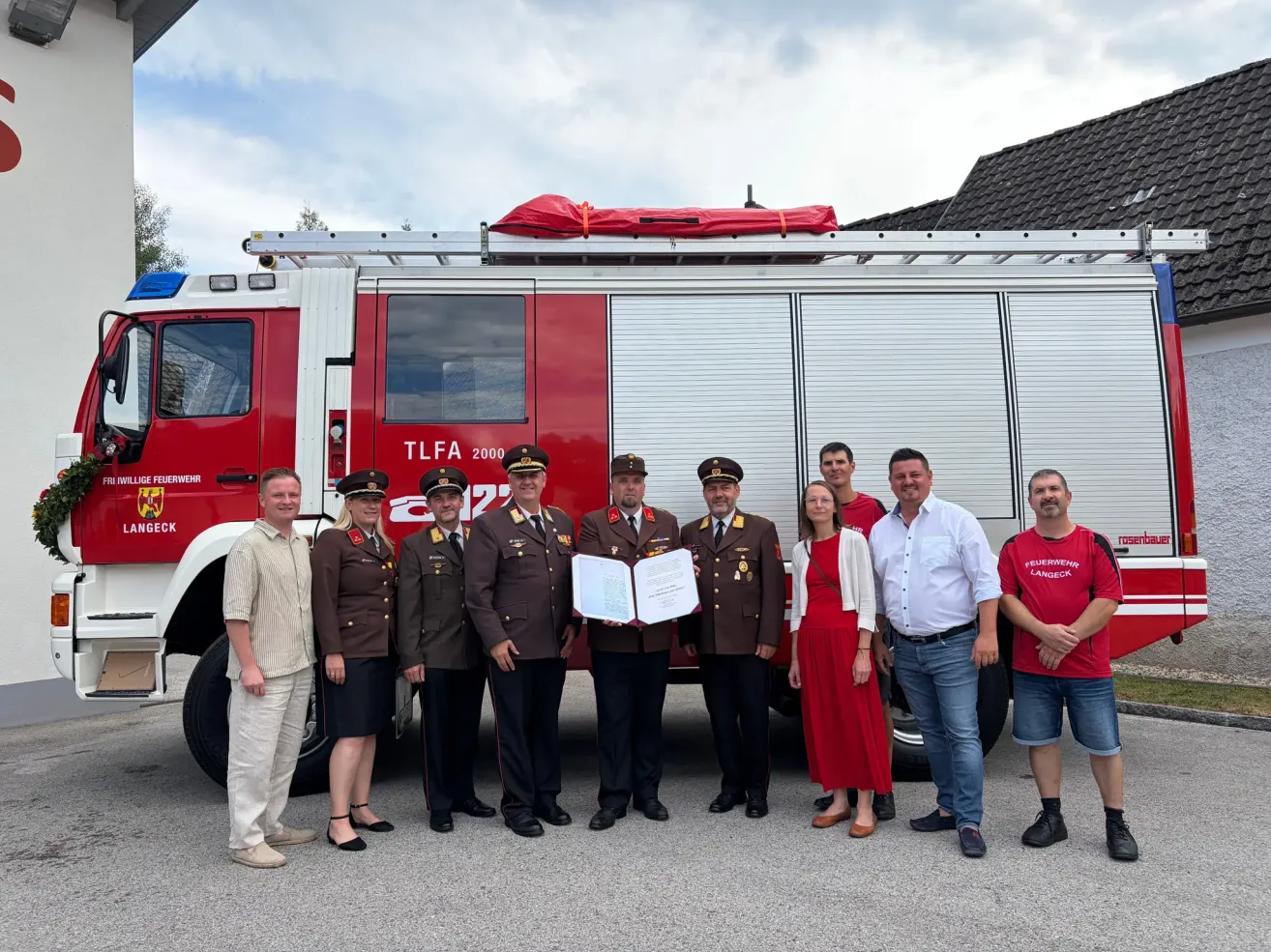 Eine Gruppe von Menschen steht vor einem roten Feuerwehrauto. Sie halten ein Zertifikat in der Hand und scheinen zu feiern. Das Feuerwehrauto hat den Text 'TLFA 2000' darauf.