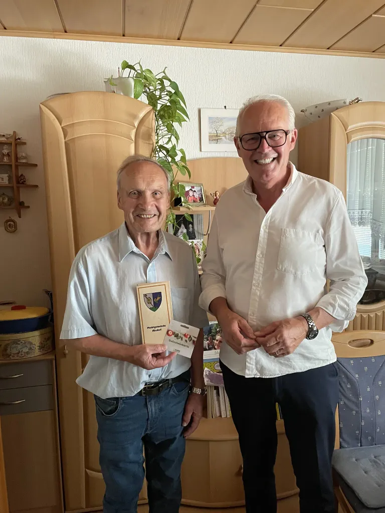 Zwei Männer stehen in einem Raum mit einem Schrank dahinter. Ein Mann hält ein Zertifikat und lächelt, während der andere Mann neben ihm steht und lächelt.