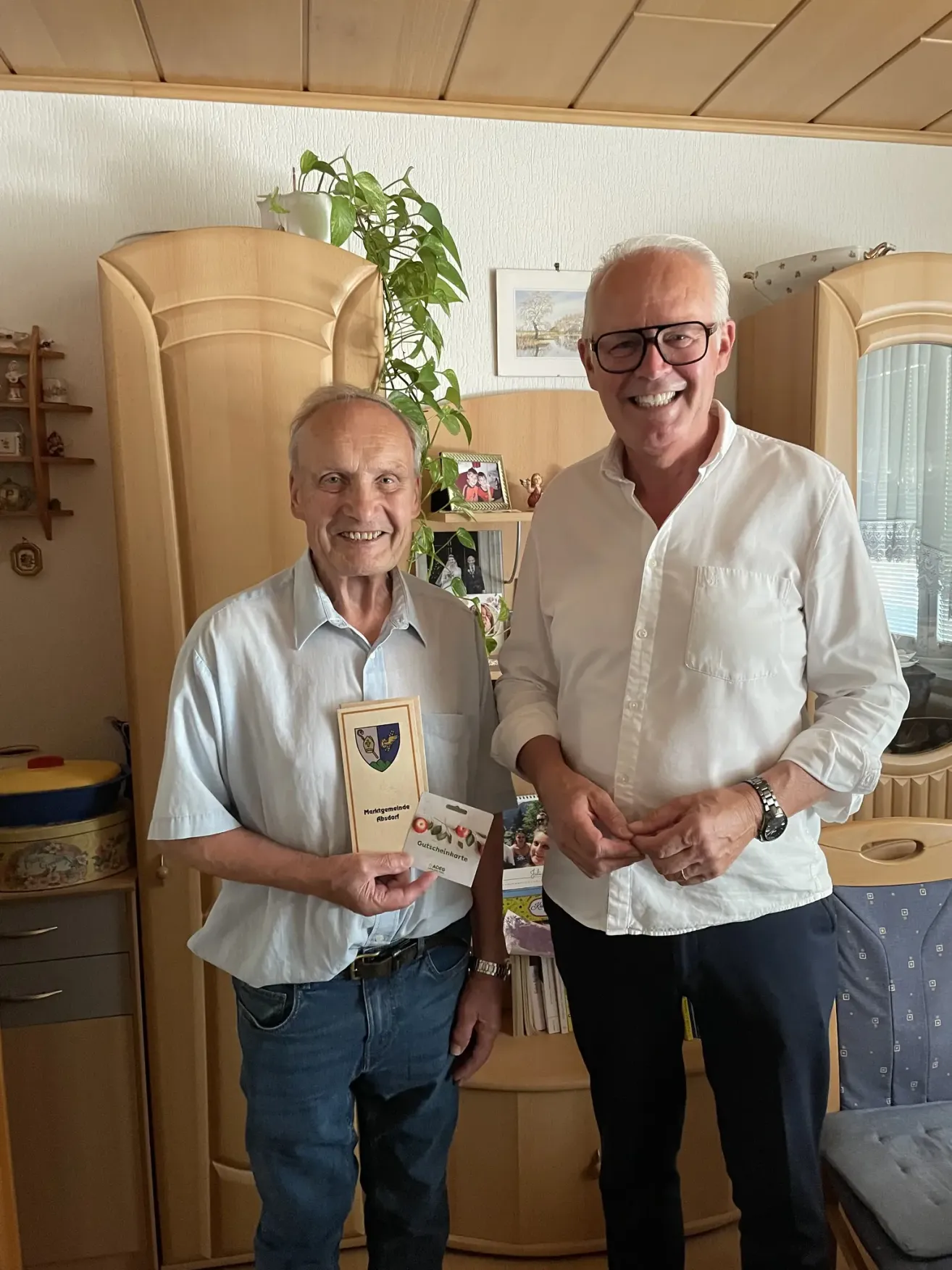 Zwei Männer stehen in einem Raum mit einem Schrank dahinter. Ein Mann hält ein Zertifikat und lächelt, während der andere Mann neben ihm steht und lächelt.