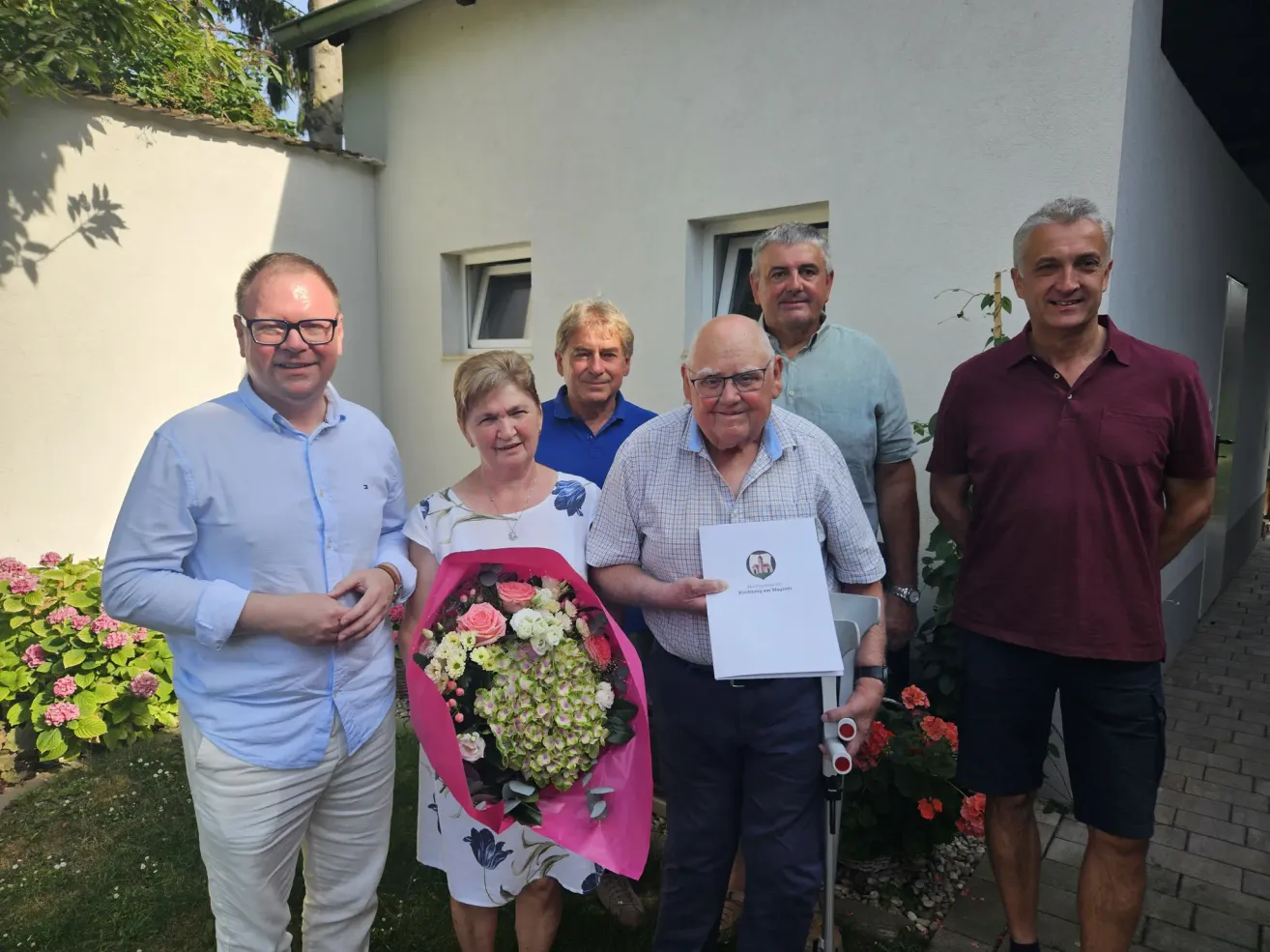 Eine Gruppe von Menschen steht vor einem Haus, einer Mann hält einen Blumenstrauß und ein anderer ein Zertifikat.