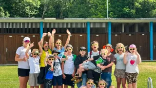 Eine Gruppe von Kindern und Erwachsenen mit Sonnenbrillen posiert für ein Foto am Schwimmbad mit einem Gebäude und Bäumen im Hintergrund.