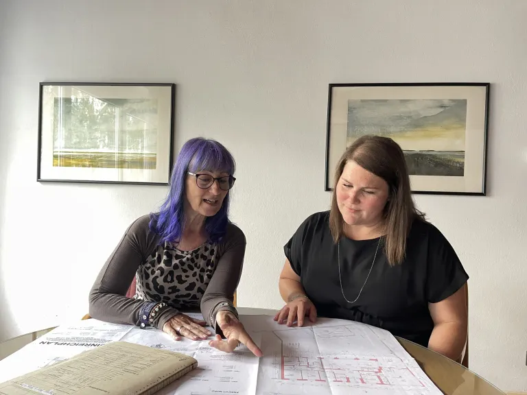 Zwei Frauen sitzen an einem Tisch mit Architekturplänen, eine Frau mit blauen Haaren zeigt auf die Pläne, während die andere Frau zusieht. Hinter ihnen hängen zwei gerahmte Gemälde an der Wand.