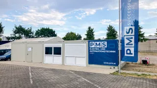 Ein modernes Gebäude mit einem blauen Banner, das 'PMS Elektro-und Automationstechnik GmbH' liest, steht auf einem Parkplatz unter einem teilweise bewölkten Himmel.