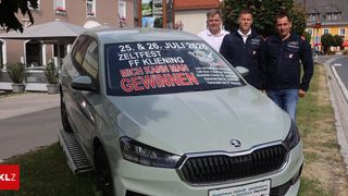 Vier Männer in Uniformen stehen neben einem Auto mit einem Schild, das 'Zeltfest FF Kleining 25. & 26. Juli 2026' und 'Mich kann man gewinnen' auf der Windschutzscheibe steht. Das Auto ist auf Gras geparkt.